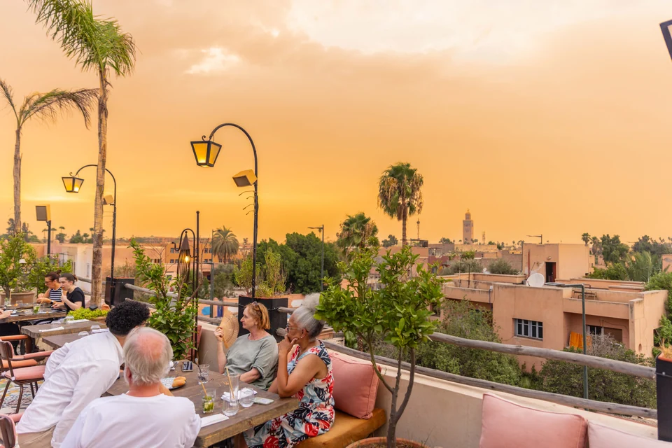 Guests dining beside terrace plants with the Koutoubia visible in the sunset skyline